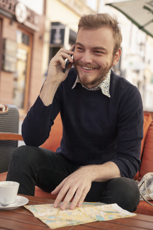 one young man - traveler or tourist talking on a phone, smiling. Sitting in a cafe outdoors, while there is a city map on a table.の写真素材