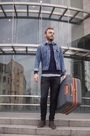 one young handsome man - traveler or tourist, carrying his suitcase in his hand. Modern building exterior, glass wall behind.の写真素材