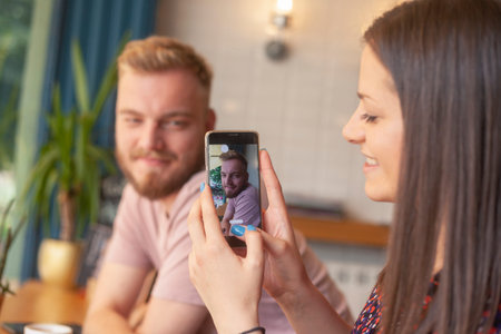 One girl talking a picture of a young man, on a smartphone. In a cafe.