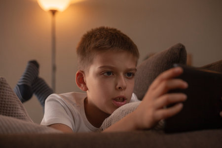 one boy, laying in bed while playing a game on his tablet. face close-up.