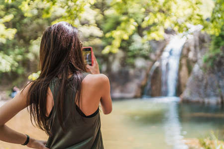 Back view of gorgeous young woman taking photos of magnificent waterfall in the forestの写真素材