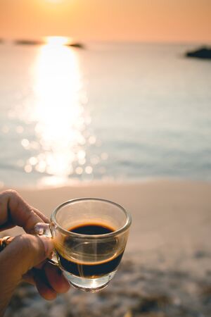 Closeup hand hold coffee cup on sand beach and view of sunset or sunrise background,peaceful,black coffee ready to drink from mug on the beach outdoor picnic.の写真素材
