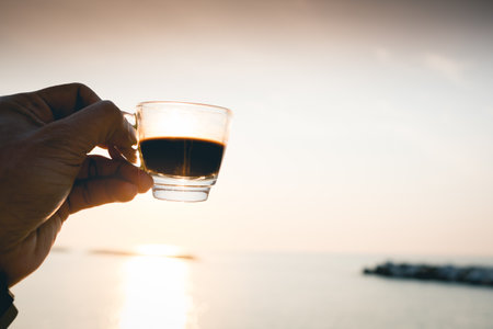 Closeup hand hold coffee cup on sand beach and view of sunset or sunrise background,peaceful,black coffee ready to drink from mug on the beach outdoor picnic.の写真素材