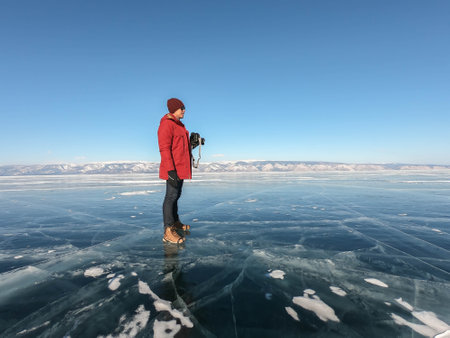 man with camera wearing bright orange winter jacket and knitted hat stands on icy frozen lake Baikalのeditorial素材