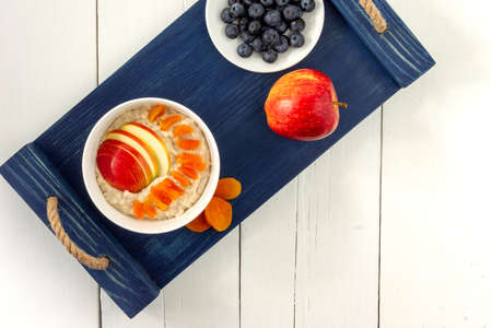 top view of porridge oatmeal bowl with blueberries and apple slices. delicious healthy breakfast. served on blue tray and white wooden tableの写真素材