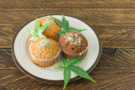 Healthy and tasty vegan and gluten free muffins topped with hemp seeds on a white plate on wooden table. Marijuana cupcake muffins with cannabis leaves.の写真素材