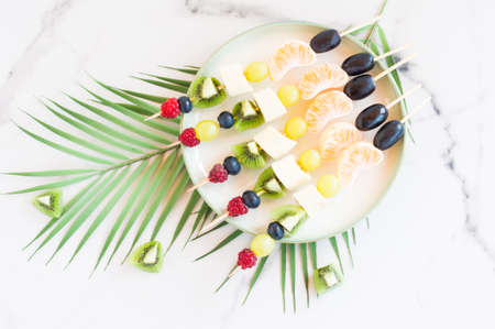 canapes of fresh fruit and berries on a plate. Marble background with palm tree branch. Festive foodの写真素材