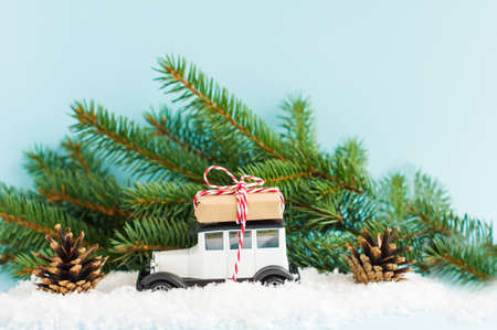 white toy retro car with a gift on the roof against the background of green spruce branches and cones in the winter snow. Christmas compositionの写真素材