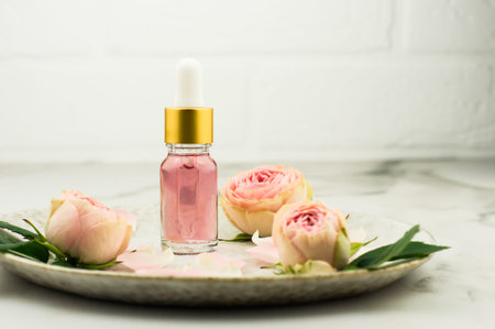 a glass bottle with a dropper pipette with essential oil of rose petals on a marble table opposite a...
