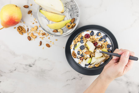 the hand of a young woman with a spoonful of granola filled during breakfast. plate with granola, berries, ripe pear. blended food, fitness, dietの写真素材