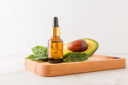 A cosmetic glass bottle of organic avocado oil stands on a wooden tray with ripe fruit and leaves...
