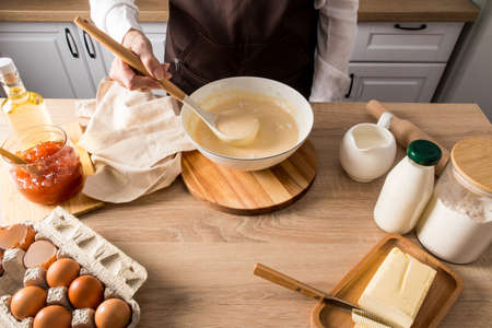 a faceless woman holds a ladle of cooked liquid pancake dough over a bowl. kitchen countertop with ingredients for further cookingの写真素材