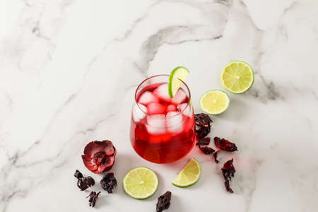 a glass of chilled hibiscus drink with ice cubes and lime wedges on a white marble table. tonic drink in the heatの写真素材