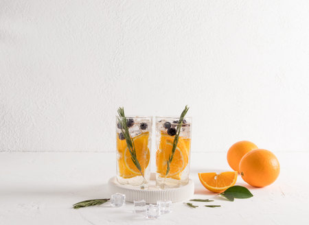 two glasses of infused water with orange and blueberries, sprigs of rosemary on a white table...