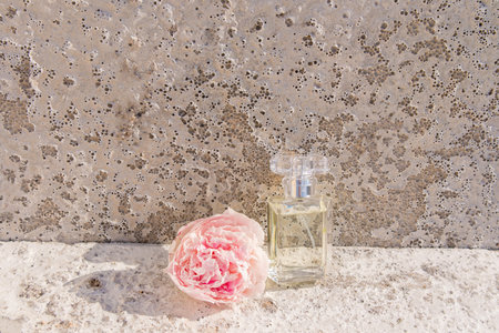 an elegant bottle of floral perfume against the background of a gray cement slab and a pink peony in...