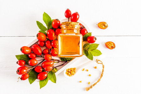a bottle with natural rosehip seed oil lies on a white tray among a pile of ripe red fruits of the...