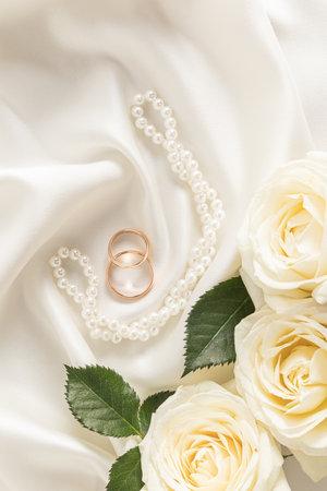 Two gold engagement rings on a satin ivory background with pearl beads and white roses. Vertical top view. layout for designの写真素材