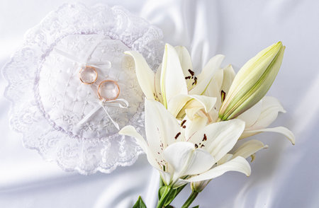 Two gold wedding rings lie on a round festive pillow. White satin background with fresh white lily flowers. top viewの写真素材