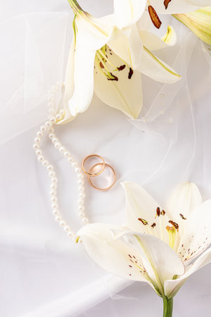 Chic vertical wedding background for design, layout, postcard, invitation. two gold wedding rings, a string of pearls and white liliesの写真素材