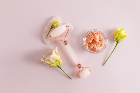 Cosmetic massager made of quartz, gua sha scraper, bath salt in a bowl on a pink background with astromeria flowers. top view. flat layoutの写真素材