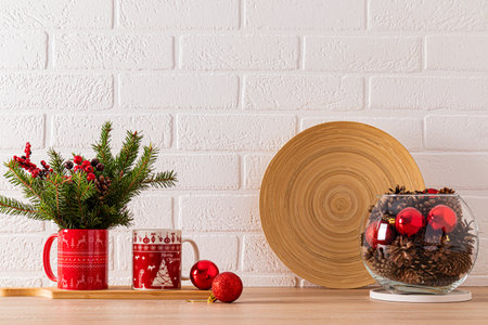 Two mugs with a Christmas pattern on the kitchen countertop with spruce branches and a round glass vase with cones and red balls. Front viewの写真素材