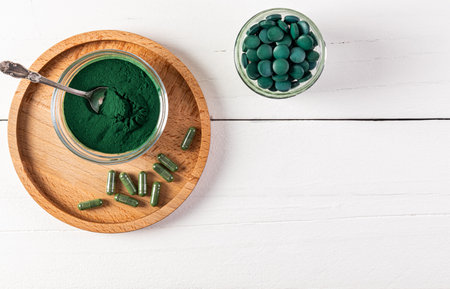 Natural Organic Green Spirulina Algae Powder In Glass Jar, Round Tablets, Capsules On Wooden Round Tray And White Background. Top viewの写真素材