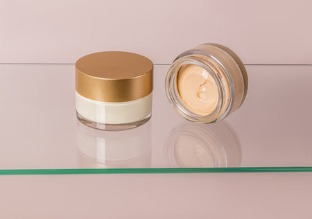 Beautiful cosmetic jar with gold lid and open jar of lying cream on glass surface. Face and body skin care. Presentation of a cosmetic productの写真素材