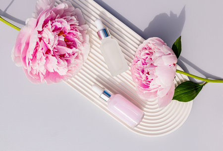 Two matte cosmetic bottles with a dropper with a serum for face and body skin care on a white embossed arch tray and peonies. top view.の写真素材