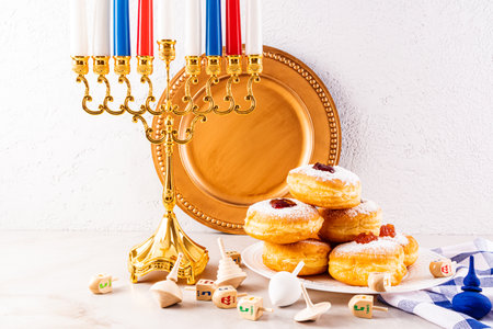 Fried donut with powdered sugar and jam on a plate, candles, minor, dreidels, gold coins on a textured wall background. The concept of the Hanukkah.の写真素材