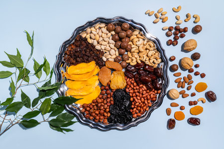 A large silver dish with a set of various dried fruits, a green branch and a scattering of dried fruits on a blue background. Upper view.の写真素材