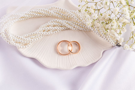 Two gold classic wedding rings are sitting on a decorative chic tray with pearl necklace and white gypsophila flowers. Delicate wedding card. layoutの写真素材