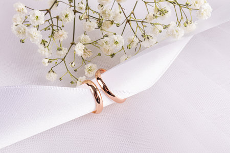 Two gold wedding rings are worn on a white satin ribbon on a white background of chic fabric with flowers. The concept of a wedding card, invitation.の写真素材