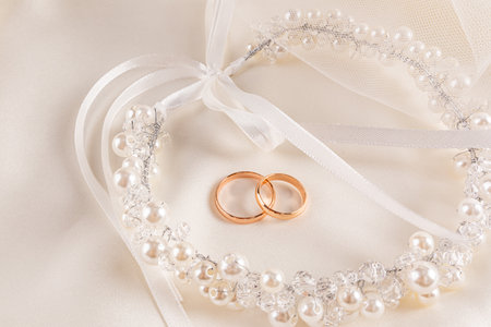 Two gold wedding rings lie on a beige satin background with a chic decoration in the bride's hairstyle with a pearl wreath with a ribbon. invitation.の写真素材
