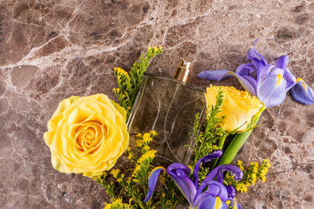 A chic bottle of perfume on a marble brown background with bright yellow and blue flowers. A blank...