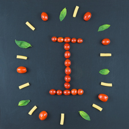 Italian food eating pattern with letter "I" composed by many red cherry tomatoes, basil leaves and macaroni isolated on blackboard backgroundの写真素材