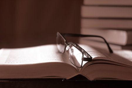 An open book with glasses lying on the pages and a stack of books in the background. Selective soft...