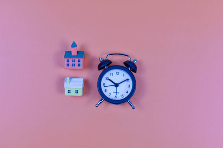 Model houses and alarm clock on pink background represent daily routines, time management, and...