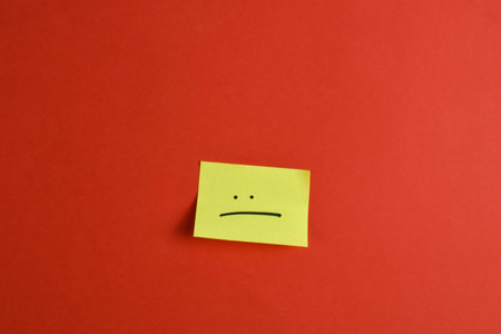Yellow sticky note with a sad face on red background, conveying a negative emotion through bold...