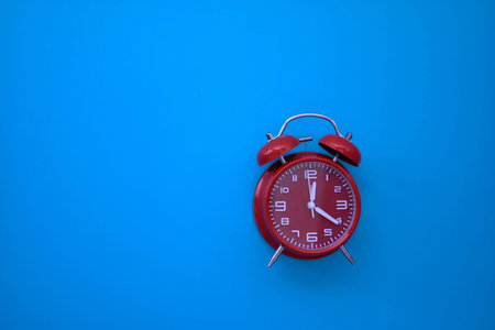 Red analog alarm clock on blue background highlights time awareness, urgency, and visual contrast in a minimalist composition.の写真素材
