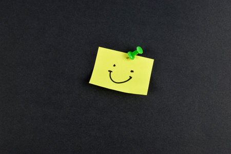Yellow sticky note with smiley face pinned by green pushpin on black background, conveying positivity through simple, bold contrast.の写真素材