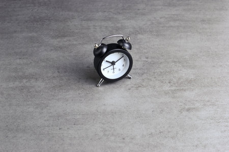 Black analog alarm clock on gray textured surface emphasizes classic timekeeping, minimalist design,...