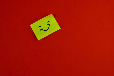 Yellow sticky note with winking smiley face on red background, conveying playful positivity through bold color contrast and minimal design.の写真素材