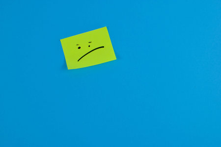 Yellow sticky note with a sad face on a bright blue background, conveying a contrasting mood through...