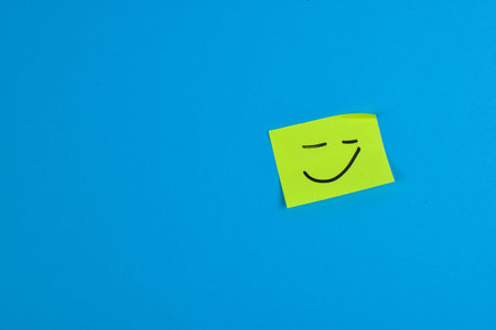 Yellow sticky note with cheerful smiley face on bright blue background, conveying positivity through bold color contrast and minimal design.の写真素材