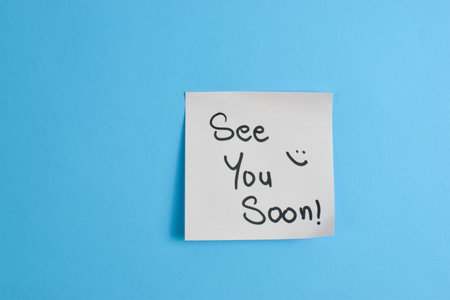 Yellow sticky note with See You Soon message on blue background, conveying warmth and greeting...