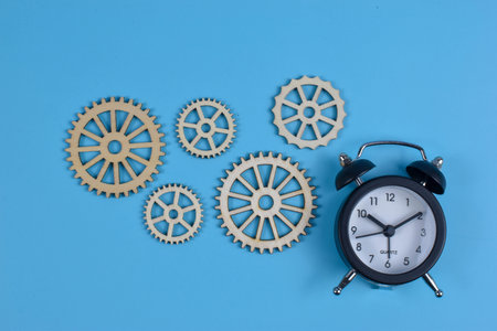 Five wooden gears and black alarm clock on blue surface show mechanical motion and time coordination in engineering or productivity themes.の写真素材