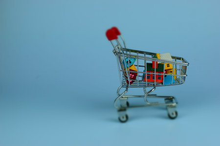 Tiny shopping cart holds colorful packages. Cart stands alone on plain blue background. Red handle adds a pop of contrast. Cart suggests retail, shopping, or delivery