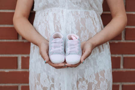 Image of a young pregnant woman posing against a brick wall in white dress holding little shoes.の写真素材