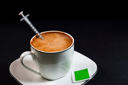 Coffee with milk in porcelain cup with syringe inside the coffee.This is a horizontal format photo taken against a black background under artificial lighting in a studio.の写真素材