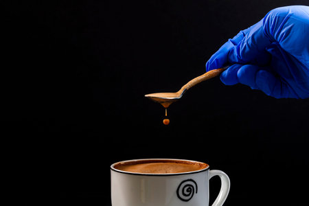 The hand of a person wearing a latex glove holds a spoon from which a drop of latte falls.This is a...
